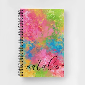 Puede incluir: Una libreta espiral colorida con un fondo de estilo acuarela en tonos de rosa, amarillo, azul y verde. La palabra "natalie" est&aacute; escrita en cursiva negra en la portada.