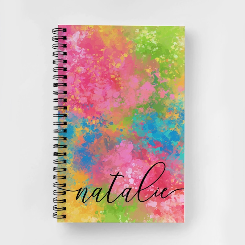 Puede incluir: Una libreta espiral colorida con un fondo de estilo acuarela en tonos de rosa, amarillo, azul y verde. La palabra "natalie" est&aacute; escrita en cursiva negra en la portada.