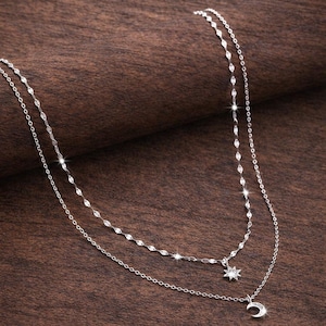Può includere: Una collana d'argento a due strati con ciondoli celesti. La catena superiore è delicata, mentre quella inferiore presenta una stella e una luna crescente. La collana è esposta su una superficie di legno marrone.