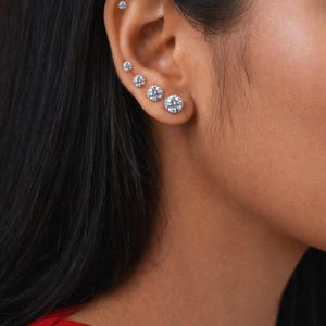 May include: A close-up of an ear adorned with six clear, round-cut diamond stud earrings. The earrings increase in size from the top of the ear to the bottom. The person's dark hair is visible on the left side of the image, and they are wearing a red top.