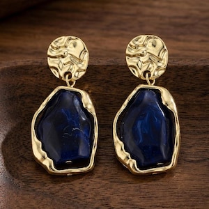 May include: A pair of gold-coloured earrings. Each earring has a textured, circular top and a large, irregular-shaped, dark blue stone pendant with a gold-coloured frame. The earrings are displayed on a wooden surface.