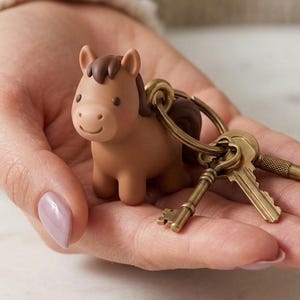 May include: A brown horse keychain with a dark brown mane and tail, attached to a brass key ring with three antique-style keys. The horse figurine is held in a hand.