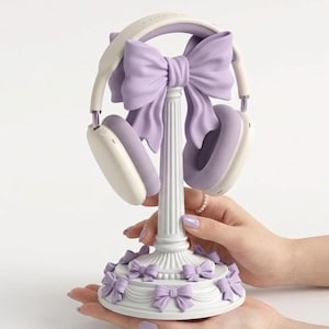 Op de afbeelding: Witte en lavendelkleurige koptelefoon op een decoratieve standaard. De standaard heeft een kolomontwerp, een grote lavendelkleurige strik en kleinere strikken rond de basis. De koptelefoon heeft witte oorschelpen en lavendelkleurige vulling.