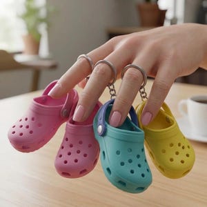 May include: Four miniature, colorful clogs keychains in pink, purple, teal, and yellow. Each clog has a silver keyring attached. The keychains are held up by a hand with pink nail polish, against a blurred background.
