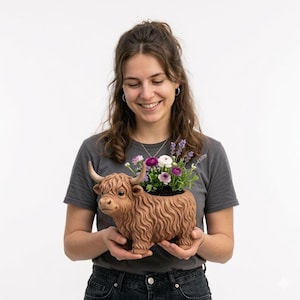 May include: A brown ceramic Highland cow planter filled with purple, white, and pink flowers. The planter has a detailed, textured design and is held by a person wearing a gray t-shirt and jeans.