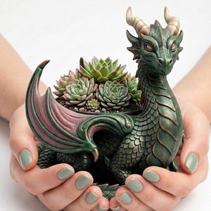 May include: A green dragon planter filled with succulents. The dragon features detailed scales, horns, and wings with pink and green accents. The planter is held in two hands with green nail polish.