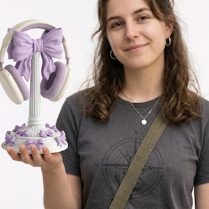 Puede incluir: Un soporte blanco para auriculares con un lazo decorativo lila y detalles de lazo a juego. El soporte sostiene un par de auriculares blancos y morados. El soporte está diseñado para sujetar auriculares y es un accesorio decorativo.