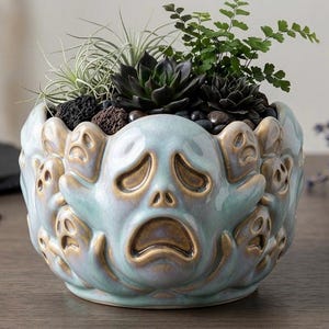 Puede incluir: Jardinera de cerámica azul claro con acabado envejecido, con un diseño de cara de fantasma en relieve. La jardinera está llena de suculentas, plantas pequeñas y piedras decorativas. La jardinera tiene aproximadamente 20 cm de diámetro.