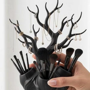 May include: Black jewelry and makeup organizer in the shape of a tree. The organizer holds necklaces, rings, earrings, and makeup brushes. The tree has multiple branches to hang jewelry and several compartments for brushes.