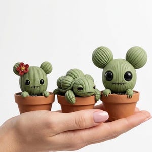 May include: Three whimsical green cactus figurines in terracotta pots. One cactus has a red flower, another resembles a turtle, and the third has large, round ears. Each has black eyes and a stitched mouth.