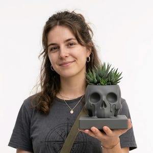 May include: A gray skull-shaped planter holding a green succulent plant. The planter has a rectangular base and a dripping design. The plant has green and white striped leaves. The planter is held by a person.