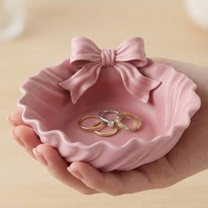 Puede incluir: Un joyero de cerámica rosa con un adorno en forma de lazo. El plato tiene un borde festoneado y contiene tres anillos. Los anillos son dorados y plateados. El plato está sostenido en una mano.