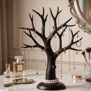 Peut inclure: Un présentoir à bijoux marron foncé en forme d'arbre, avec des branches pour accrocher colliers, bagues et boucles d'oreilles. L'arbre est sur une surface blanche, avec un miroir, des flacons de parfum et des pinceaux de maquillage en arrière-plan.