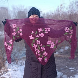 Pu&ograve; includere: Uno scialle lavorato a maglia bordeaux con ricami floreali rosa. Lo scialle &egrave; tenuto in alto, mostrando l'intricato design floreale. Lo sfondo &egrave; una scena invernale con neve e alberi.