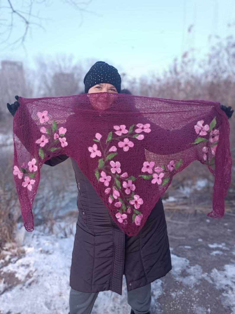 Pu&ograve; includere: Uno scialle lavorato a maglia bordeaux con ricami floreali rosa. Lo scialle &egrave; tenuto in alto, mostrando l'intricato design floreale. Lo sfondo &egrave; una scena invernale con neve e alberi.
