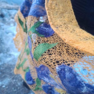 Pu&ograve; includere: Primo piano di una sciarpa fatta a mano con trama aperta. La sciarpa presenta una vivace tavolozza di colori, con una base giallo dorato e accenti blu, verdi e gialli. Il motivo floreale crea un accessorio unico e artistico.