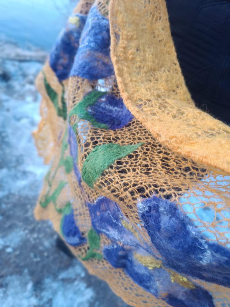 Pu&ograve; includere: Primo piano di una sciarpa fatta a mano con trama aperta. La sciarpa presenta una vivace tavolozza di colori, con una base giallo dorato e accenti blu, verdi e gialli. Il motivo floreale crea un accessorio unico e artistico.