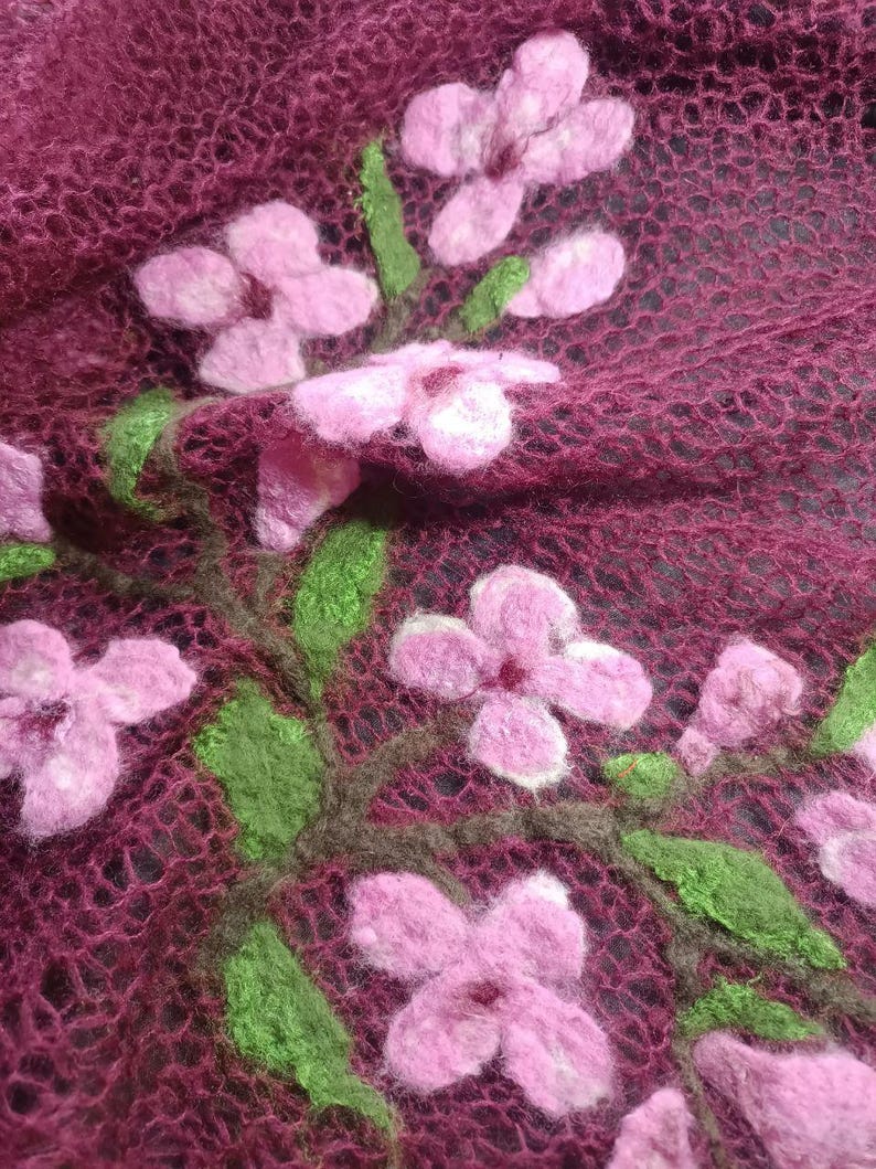 Pu&ograve; includere: Primo piano di un tessuto bordeaux a trama aperta, ornato da fiori rosa infeltriti e foglie verdi. I fiori, con centri bianchi, sono disposti in un motivo floreale. Il tessuto ha un aspetto delicato e arioso, che suggerisce una sciarpa o uno scialle.