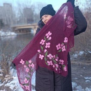 Pu&ograve; includere: Uno scialle lavorato a maglia bordeaux con ricami floreali rosa. Lo scialle &egrave; esposto, evidenziando il motivo floreale dettagliato e gli steli verdi. Lo sfondo presenta una persona con cappello e cappotto neri, sullo sfondo di un paesaggio innevato.