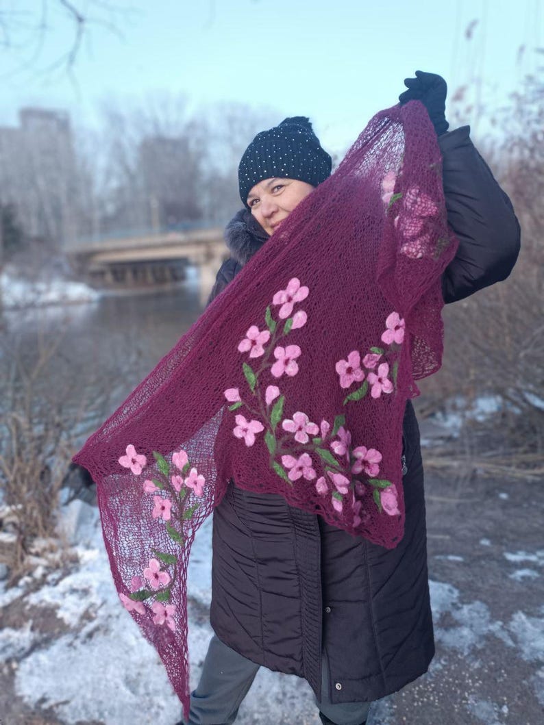 Pu&ograve; includere: Uno scialle lavorato a maglia bordeaux con ricami floreali rosa. Lo scialle &egrave; esposto, evidenziando il motivo floreale dettagliato e gli steli verdi. Lo sfondo presenta una persona con cappello e cappotto neri, sullo sfondo di un paesaggio innevato.