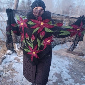Pu&ograve; includere: Uno scialle nero con un motivo floreale con grandi fiori rossi con centri gialli e foglie verdi. Lo scialle &egrave; tenuto in alto, mostrando gli intricati dettagli dei fiori ricamati. La persona indossa un cappello e guanti neri.
