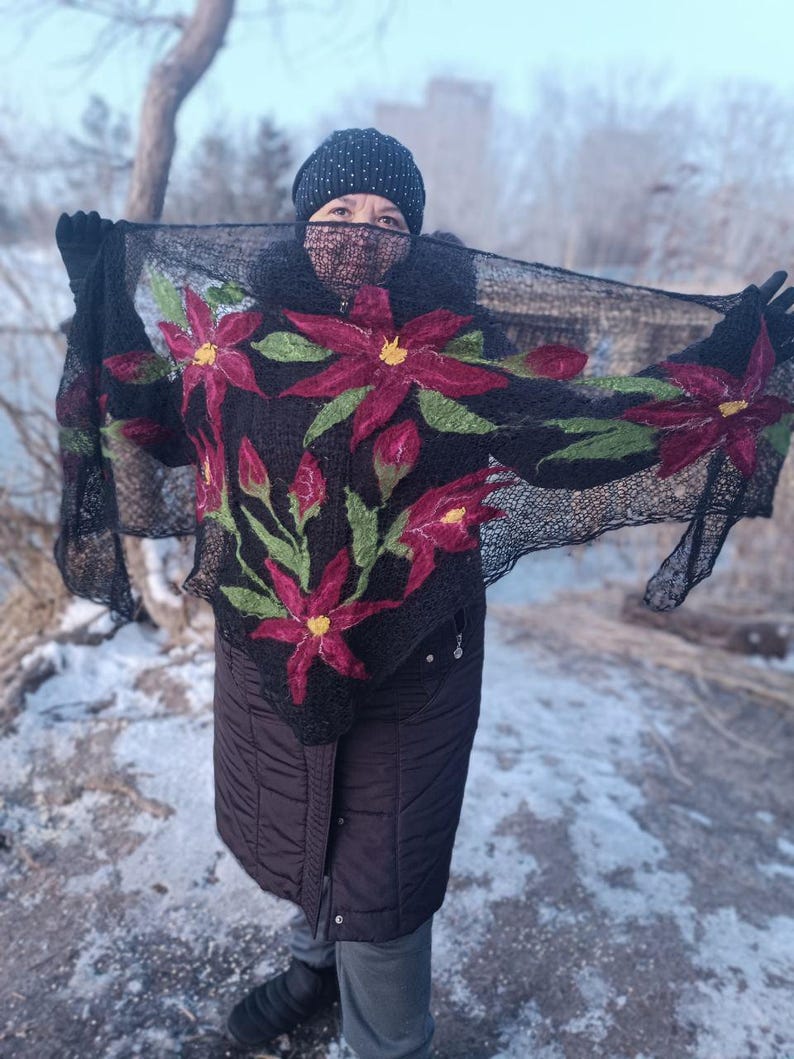 Pu&ograve; includere: Uno scialle nero con un motivo floreale con grandi fiori rossi con centri gialli e foglie verdi. Lo scialle &egrave; tenuto in alto, mostrando gli intricati dettagli dei fiori ricamati. La persona indossa un cappello e guanti neri.