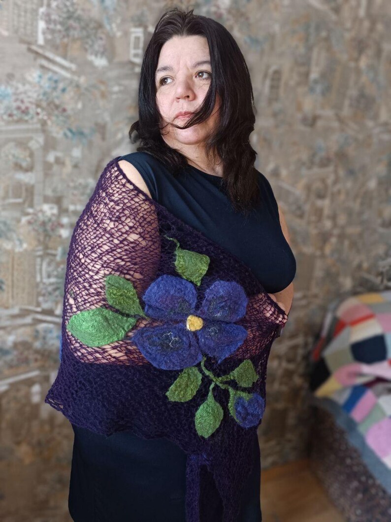 Pu&ograve; includere: Uno scialle viola scuro con un grande fiore blu ricamato e foglie verdi. Lo scialle ha una trama a rete ed &egrave; drappeggiato sulle spalle. Il fiore ha un centro giallo.