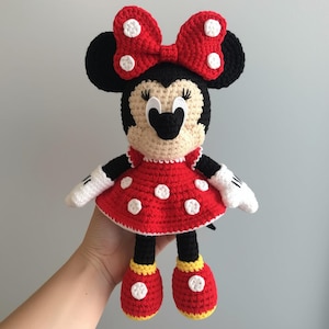 May include: A handmade crochet doll of a cartoon character. The doll has a black head with large ears, a red bow with white polka dots, and a red dress with white polka dots. The doll's shoes are red with yellow soles.