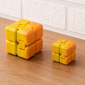 Könnte beinhalten: Zwei gelb-orangefarbene Fidget-Würfel liegen auf einer Holzoberfläche. Der größere Würfel besteht aus acht verbundenen Blöcken, während der kleinere Würfel ein ähnliches Design hat. Die Würfel haben einen Farbverlauf von Gelb nach Orange.