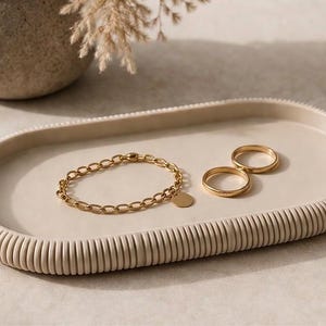 Könnte beinhalten: Ein beigefarbenes ovales Tablett mit geripptem Rand enthält ein goldfarbenes Kettenarmband mit einem kreisförmigen Anhänger und zwei goldfarbenen Ringen. Im Hintergrund befindet sich eine beige Vase mit getrockneten Pflanzen. Die Artikel sind auf einer hellen Oberfläche angeordnet.