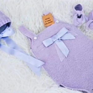 Könnte beinhalten: Ein Set aus lavendelfarbener Strickkleidung für Babys, bestehend aus einer Mütze mit Bändern, einem Strampler mit Schleife und passenden Babyschuhen. Das Set wird auf einer weißen, flauschigen Oberfläche präsentiert. Ein kleines Etikett ist am Strampler befestigt.