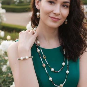 Op de afbeelding: Een vrouw draagt een groene jurk en een parelsieradenset, inclusief een ketting, oorbellen en armband. De sieraden hebben grote, ronde parels met kleurrijke accenten. De achtergrond toont witte bloemen en groen.