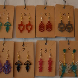 May include: Nine pairs of earrings are displayed on brown tags. The earrings showcase diverse designs, such as geometric shapes, stylized arrows, and fairy wings, in colours like green, red, purple, and gold. Each pair hangs from a silver or gold hook.