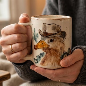 May include: A ceramic mug with a speckled off-white finish featuring a hand-painted illustration of a duck wearing a brown hat. The duck has a yellow-orange beak and brown feathers, with small blue-green leaf accents painted around the figure.