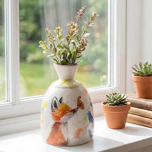 Peut inclure: Un vase en céramique décoratif avec un renard et un papillon, rempli de verdure, sur un rebord de fenêtre. Le vase est blanc avec des éclaboussures de peinture colorées. Deux petits pots en terre cuite avec des succulentes sont également sur le rebord.