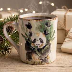 Op de afbeelding: Een keramische mok met een rustieke, verweerde bruine rand, voorzien van een aquarelillustratie van een panda die bamboestengels eet. De mok is gevuld met een donkere vloeistof en heeft een handvat aan de linkerkant.