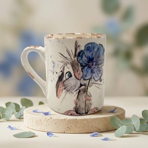 Może przedstawiać: Ceramiczny kubek o kremowej podstawie i rustykalnym brązowym rancie. Kubek ozdobiony jest ręcznie malowaną ilustracją małego, szarego, futrzanego stworzenia trzymającego duży, żywy niebieski kwiat. Wzór zawiera delikatne niebieskie rozpryski farby.