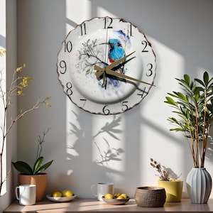 May include: White ceramic wall clock with a blue bird illustration. The clock has black numbers and gold-colored hands. The clock's edge has a wavy, textured design. The clock is mounted on a white wall.