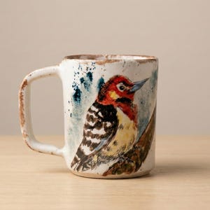 May include: A white ceramic mug with a hand-painted illustration of a woodpecker perched on a branch. The bird features a red head, black and white striped wings, and a yellow chest. The mug has a rustic, textured finish with brown edges.