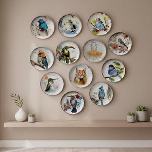 以下が含まれることがあります： 水彩画の鳥と猫のデザインが施された、12枚の装飾用プレートのコレクション。プレートは丸く、金属の縁が施され、ベージュの壁に配置されています。プレートには、さまざまな鳥と猫が描かれています。