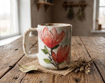 May include: A ceramic mug with a speckled off-white glaze featuring a hand-painted illustration of two large pinkish-red magnolia flowers with green leaves and dark outlines. The mug has a thick, sturdy handle and a slightly irregular, organic shape.