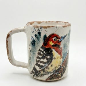 May include: A ceramic mug with a speckled off-white finish and a brown rim. The exterior features a hand-painted illustration of a woodpecker with a red head, black and white striped wings, and a yellow chest, perched on a brown branch.