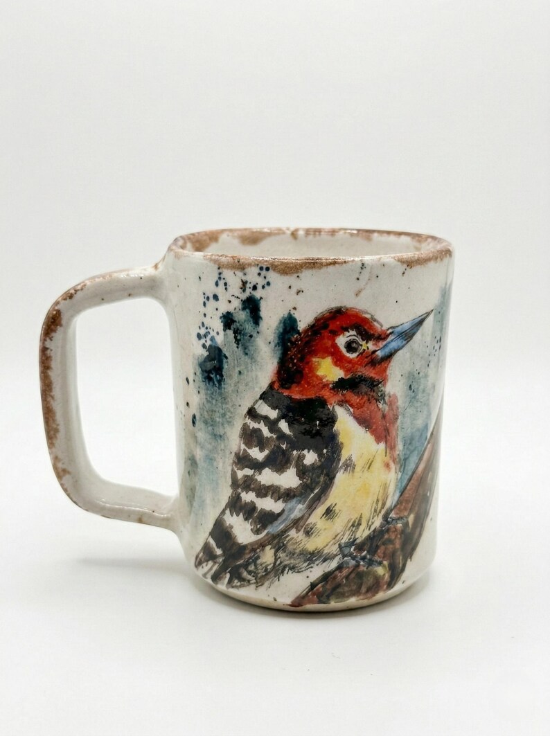 May include: A ceramic mug with a speckled off-white finish and a brown rim. The exterior features a hand-painted illustration of a woodpecker with a red head, black and white striped wings, and a yellow chest, perched on a brown branch.