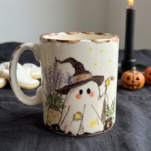 May include: A cream-colored ceramic mug featuring a hand-painted illustration of a ghost wearing a witch hat and holding a star wand. The design includes lavender sprigs, small yellow stars, and a patch on the ghost, with a brown rim accent.