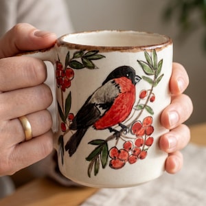 May include: A cream-colored ceramic mug featuring a hand-painted bullfinch perched on a branch with red berries and green leaves. The mug has a textured, speckled finish and a brown rim along the top edge, with a sturdy handle on the side.