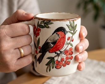 May include: A cream-colored ceramic mug featuring a hand-painted bullfinch perched on a branch with red berries and green leaves. The mug has a textured, speckled finish and a brown rim along the top edge, with a sturdy handle on the side.