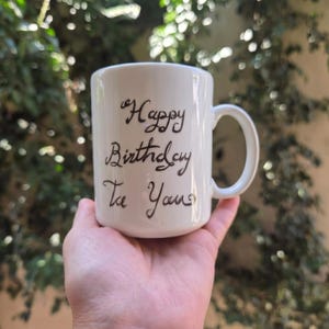May include: A white ceramic mug with a rounded handle featuring the handwritten black text Happy Birthday To You on the front surface. The mug has a smooth, glossy finish and a cylindrical shape, designed for holding hot or cold beverages.
