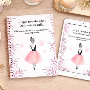 Puede incluir: Un diario con espiral y una tableta que muestran una ilustración de bailarina con texto en español. El diario y la tableta están rodeados por una taza de café, flores y artículos de papelería sobre una superficie de madera clara. El texto dice 'Lo que no sabes de ti'.