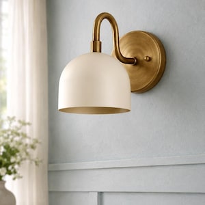 May include: A wall-mounted light fitting featuring a curved brass arm and a circular brass backplate. The lamp includes a smooth, dome-shaped off-white shade that directs light downwards. The metal components have a brushed, matte brass finish.