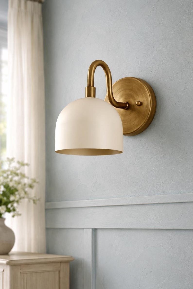 May include: A wall-mounted light fitting featuring a curved brass arm and a circular brass backplate. The lamp includes a smooth, dome-shaped off-white shade that directs light downwards. The metal components have a brushed, matte brass finish.